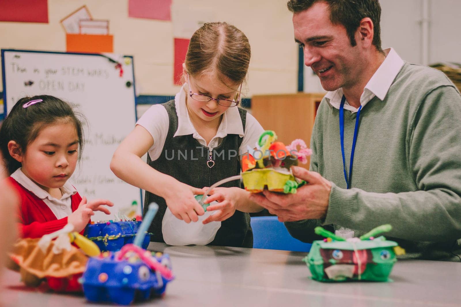Primary school teacher is helping two of his students with a STEM project. They are building something using recycled items and crafts equipment.