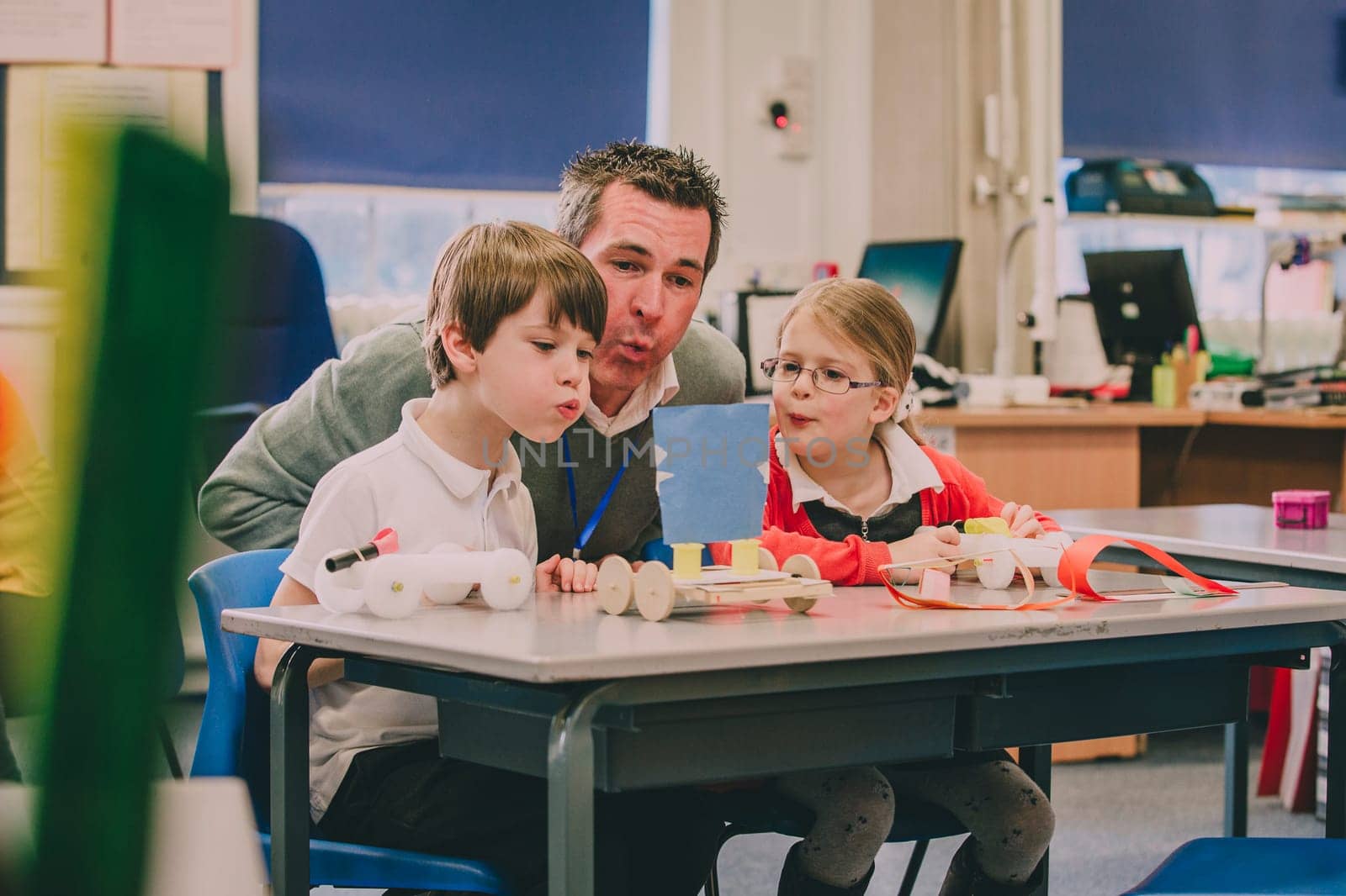 Teacher is sitting with two of his primary school students in the classroom, helping them test their physics project. They are creating air by blowing with their mouths, to move their toy vehicles.