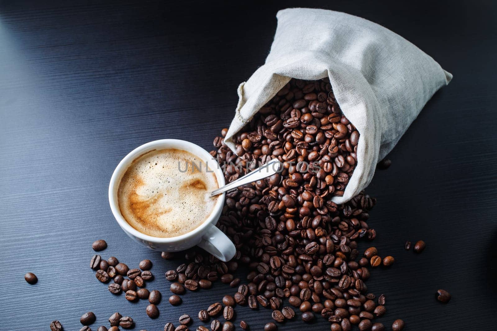 Cup of espresso and coffee beans scattered from a canvas bag on black wooden table background.