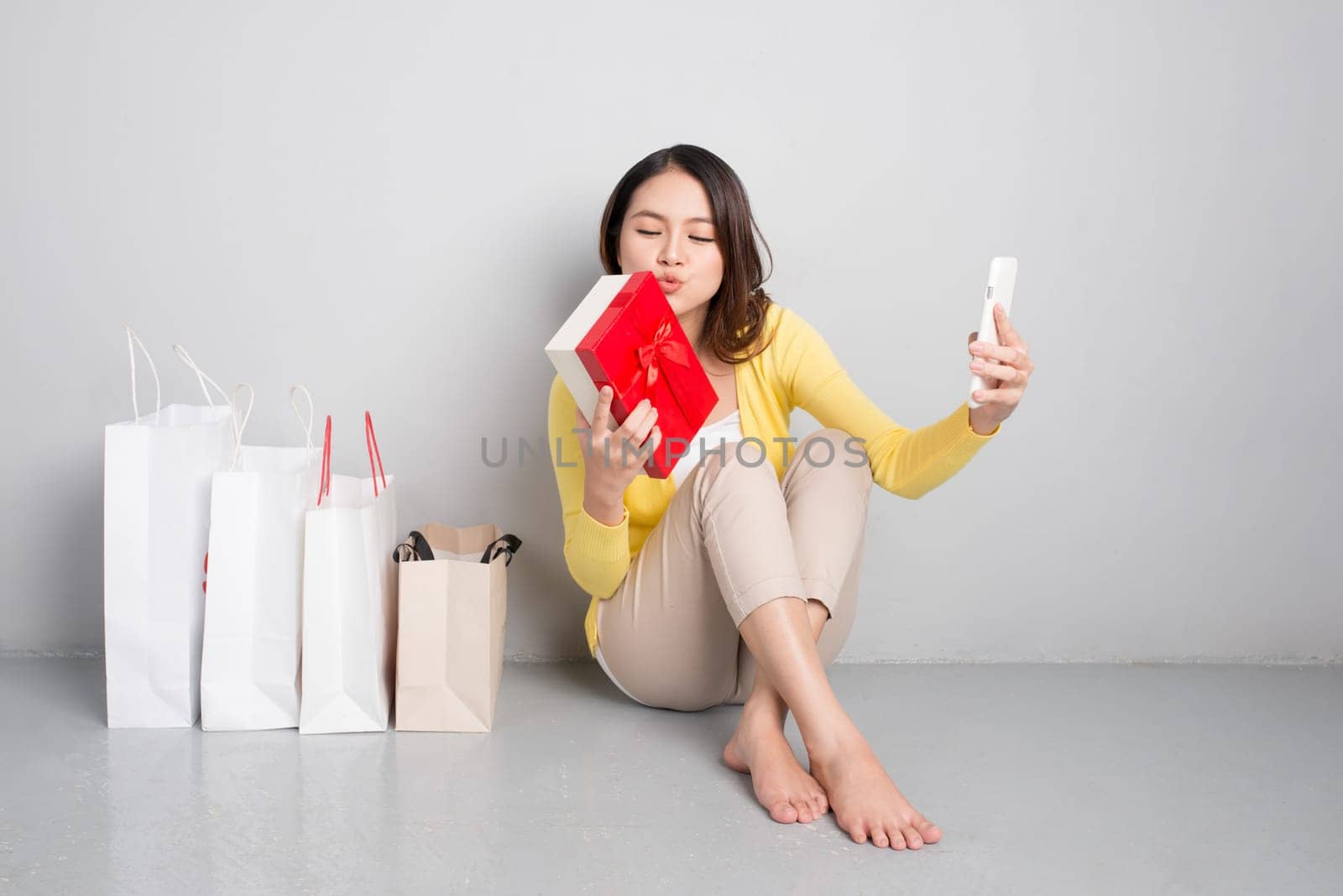 Young asian woman sitting besides row of shopping bags holding red gift box taking sellfie