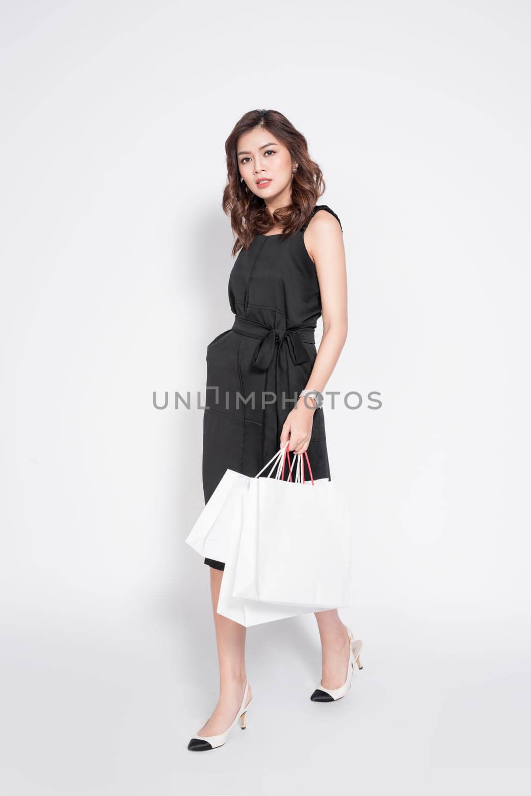 Beautiful asian woman wearing black dress with shopping bag standing over white.