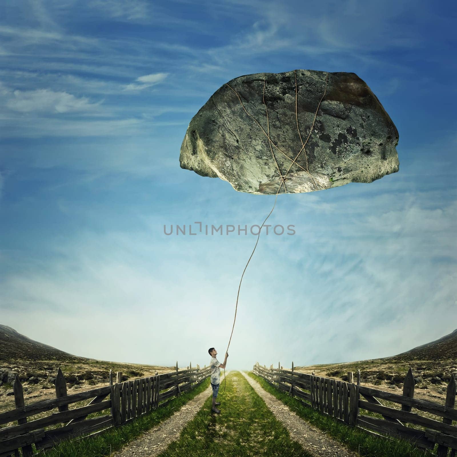 Surrealistic image as a young boy stand on a country road, holding a rope bound around a huge stone as a playing kite. Life pressure, stress and hard determination concept.