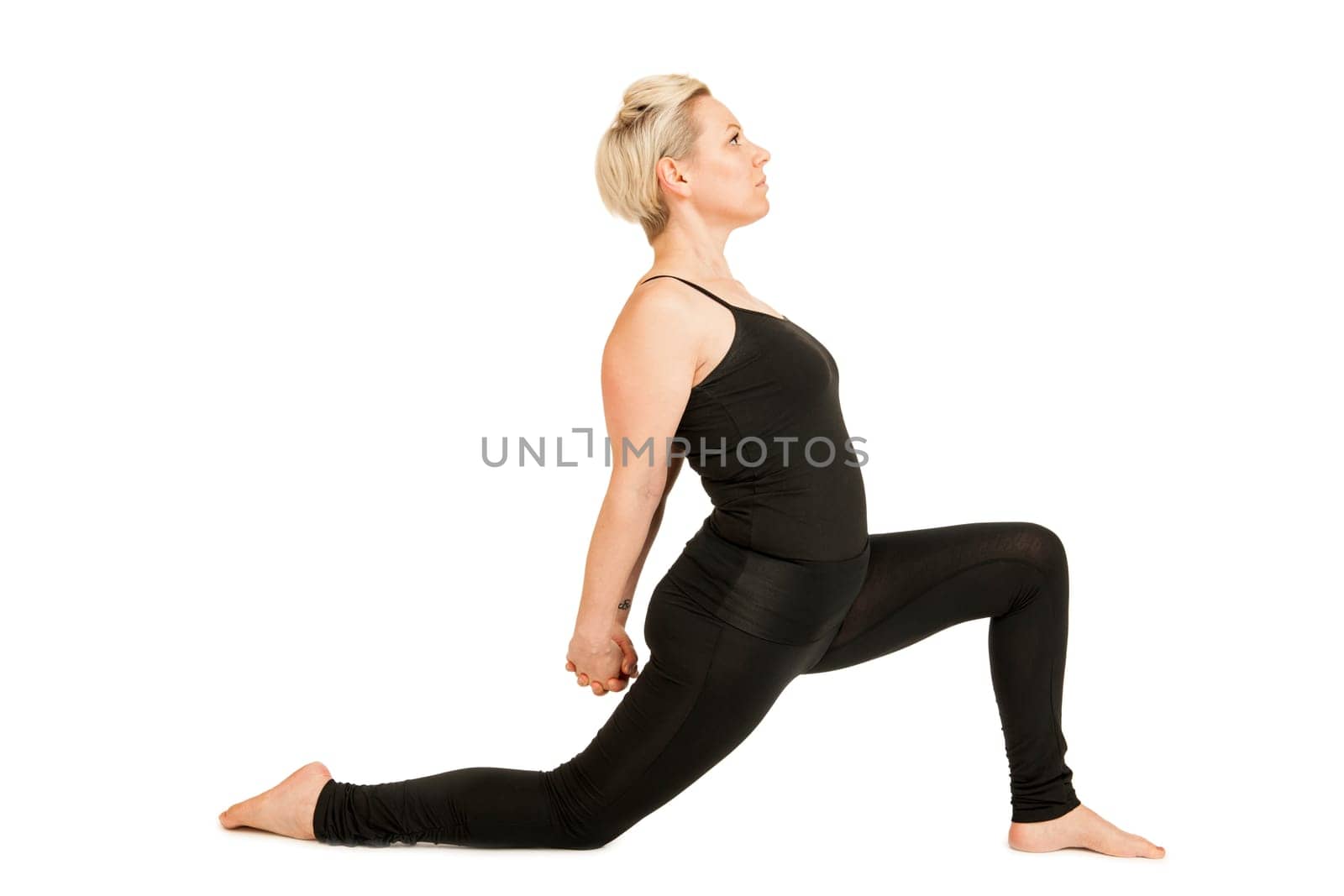 Lateral full-body view of a young woman on a white background in a black sports dress showing a variation of the yoga exercise hero position (Anjaneyasana) with a deep lunge.
