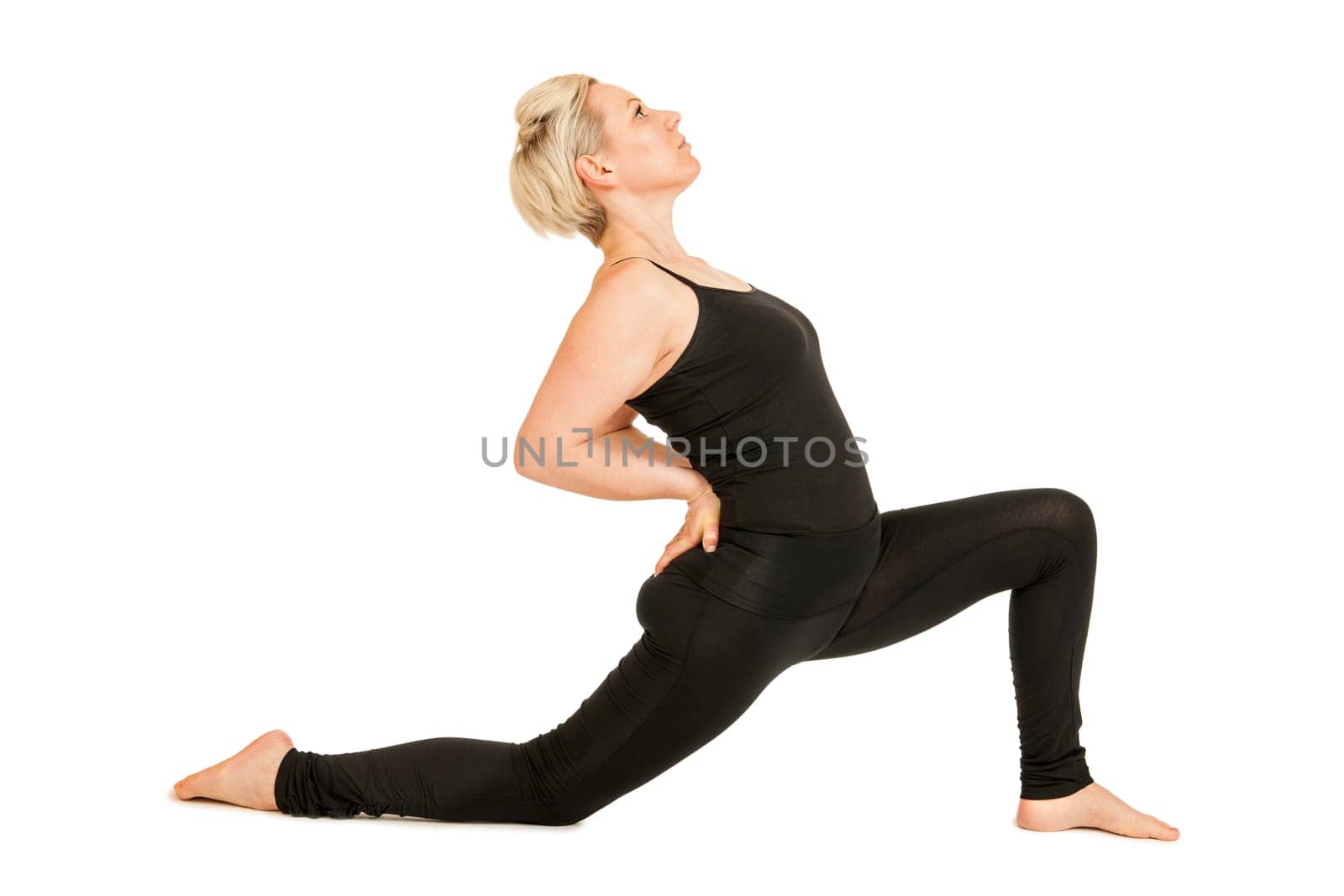 Lateral full-body view of a young woman on a white background in a black sports dress showing a variation of the yoga exercise hero position (Anjaneyasana) with a deep lunge.