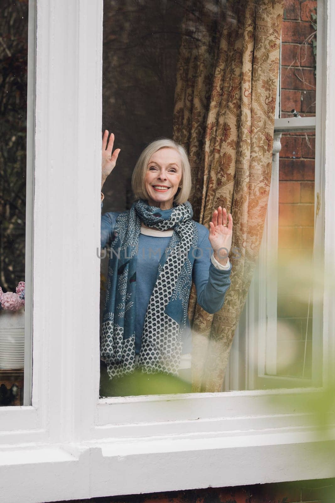 Senior woman is waving goodbye from a window in her home.