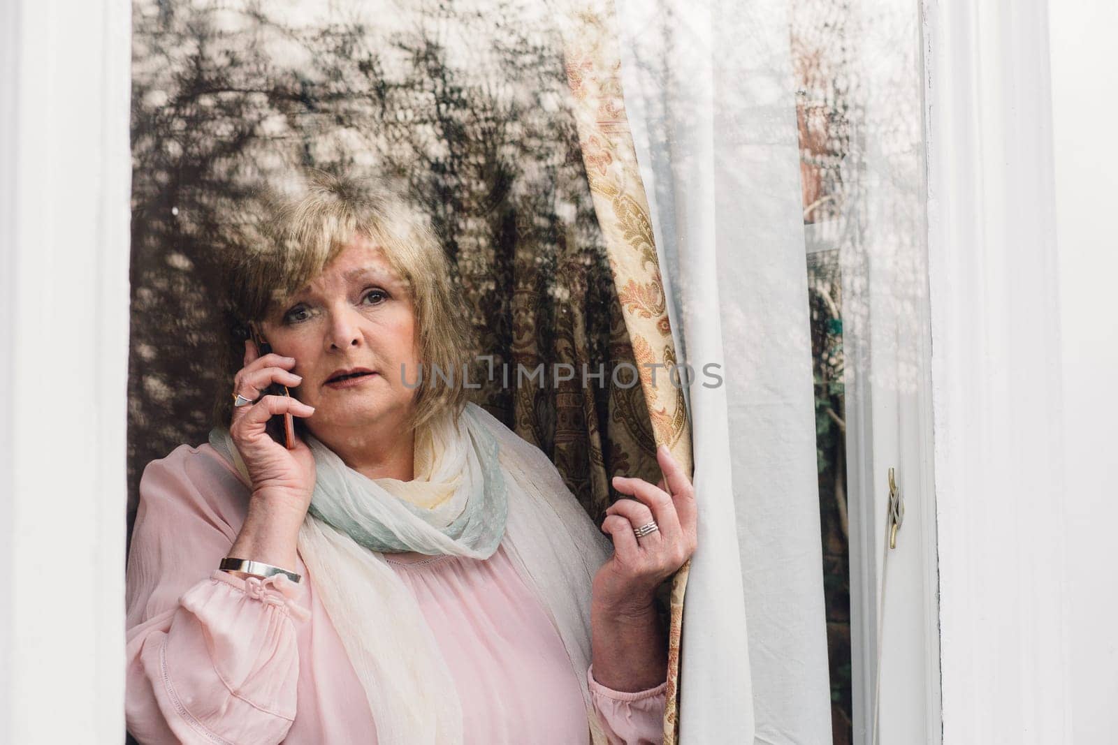 Senior woman is reporting a neighborhood crime to the police on the phone. She is looking out her window.