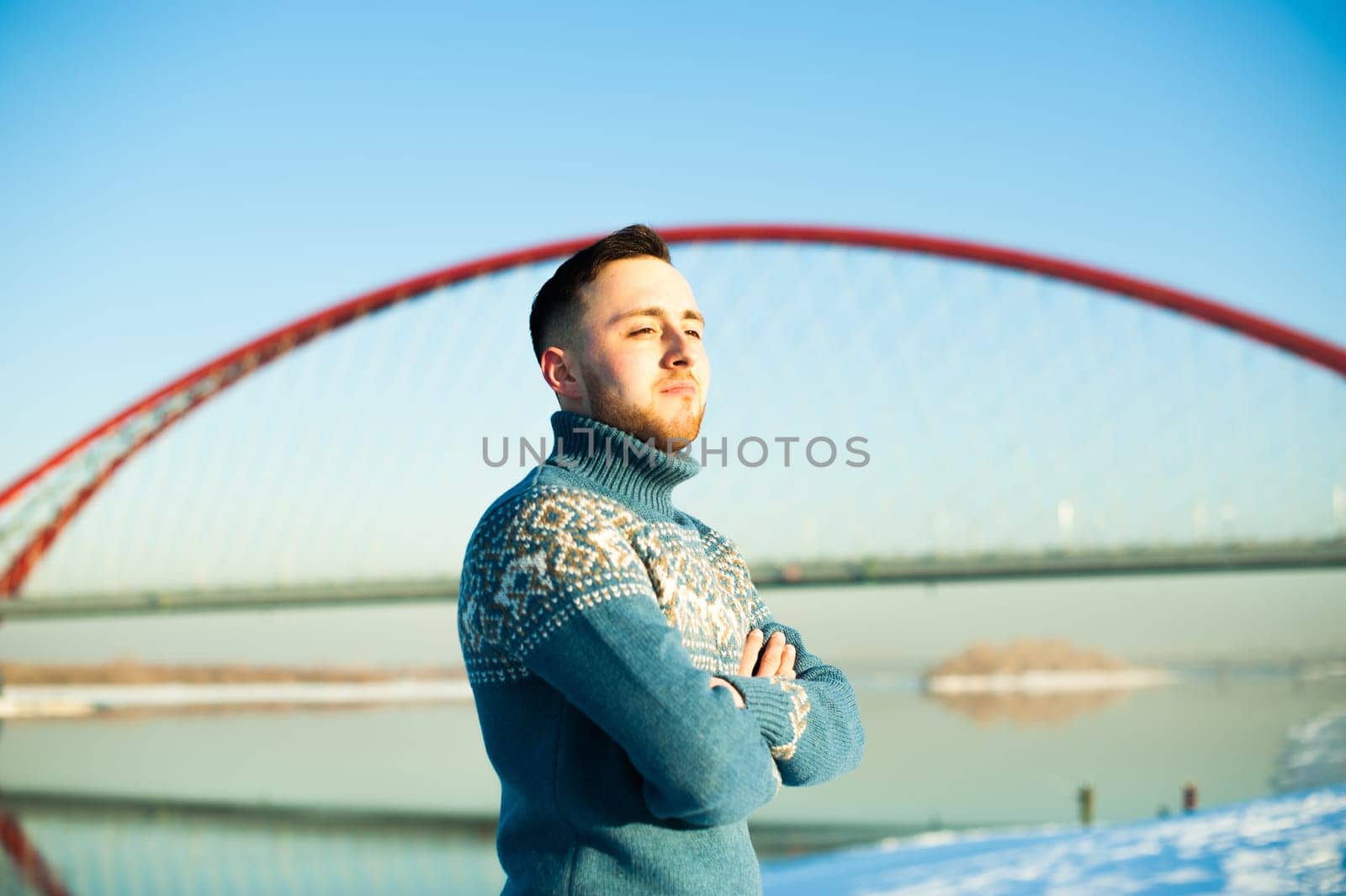 man in warm sweater with a beard is walking on the street in the winter in a warm sunny day by the river on a background of red bridge