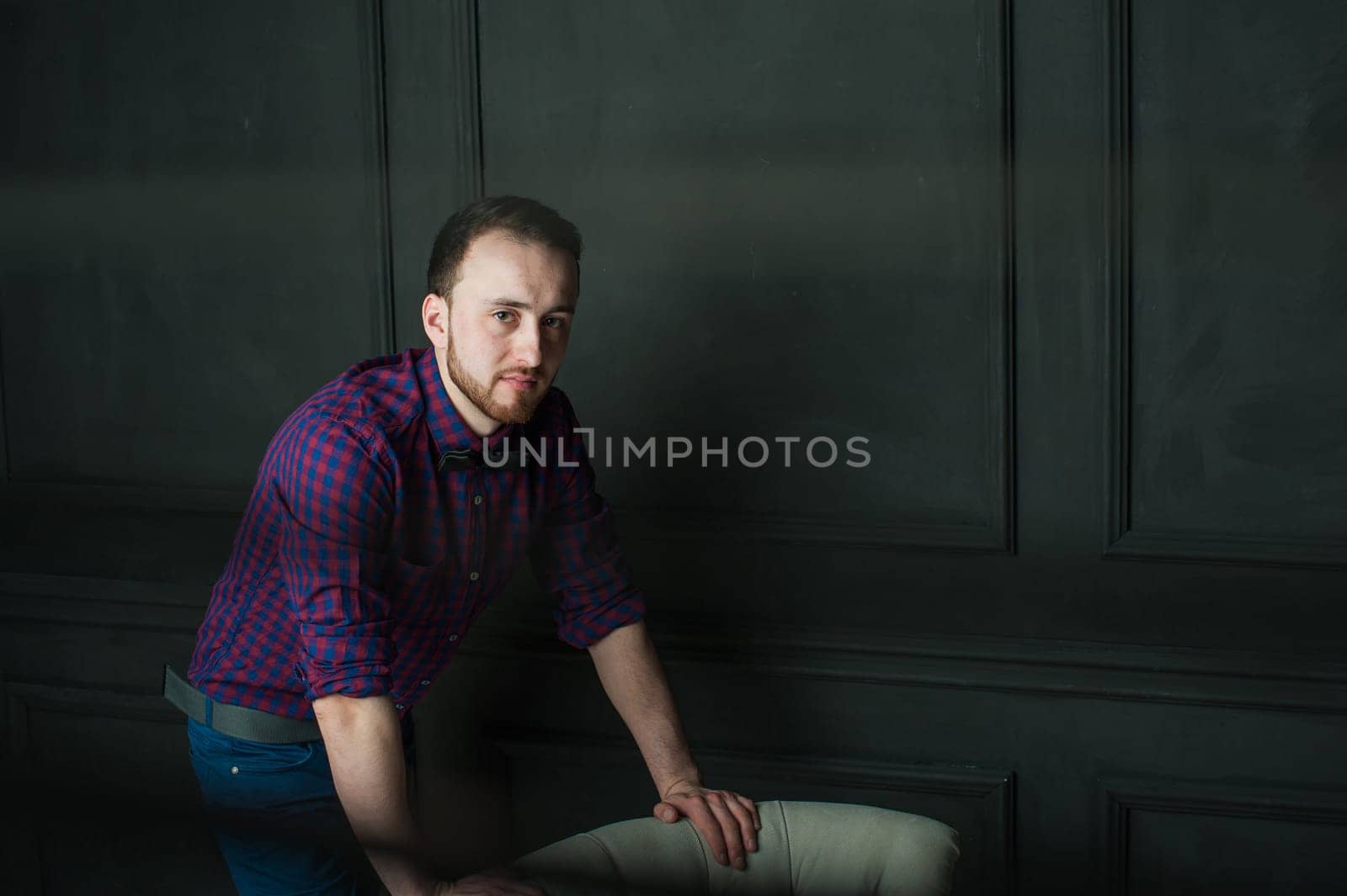 studio portrait of a young man on a dark wall background in a burgundy plaid shirt and blue jeans. hipster man, dressed stylishly, with a beard, in a room with an armchair