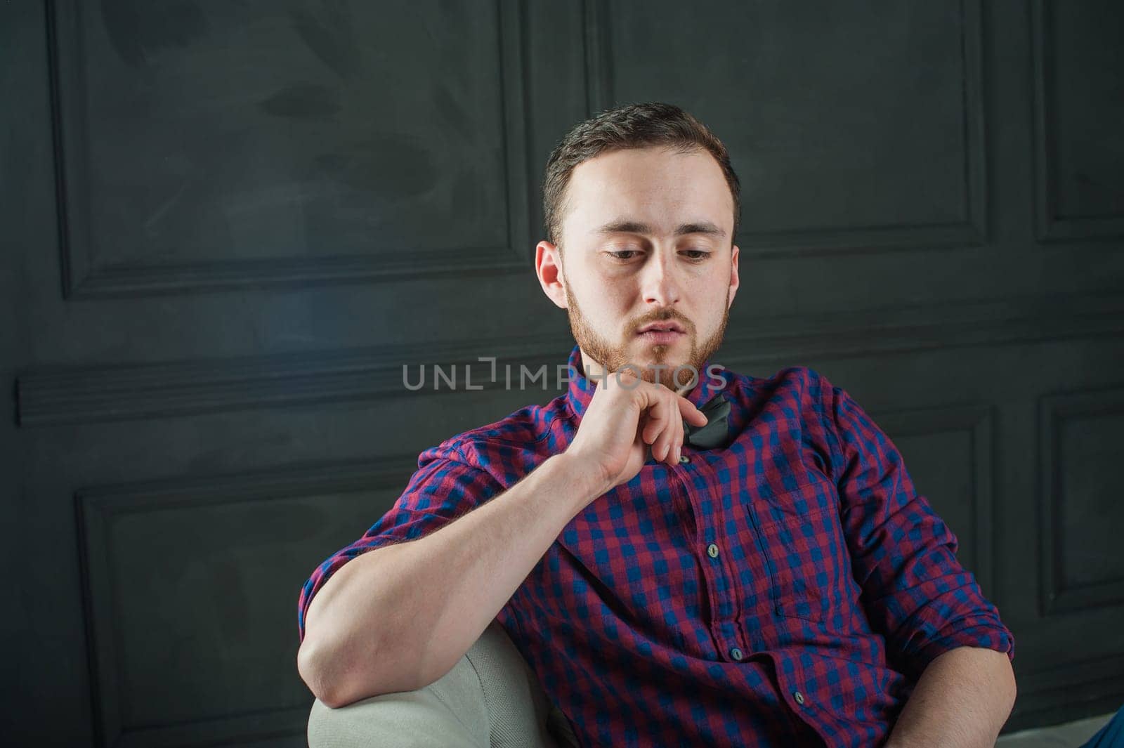 studio portrait of a young man on a dark wall background in a burgundy plaid shirt and blue jeans. hipster man, dressed stylishly, with a beard, in a room with an armchair