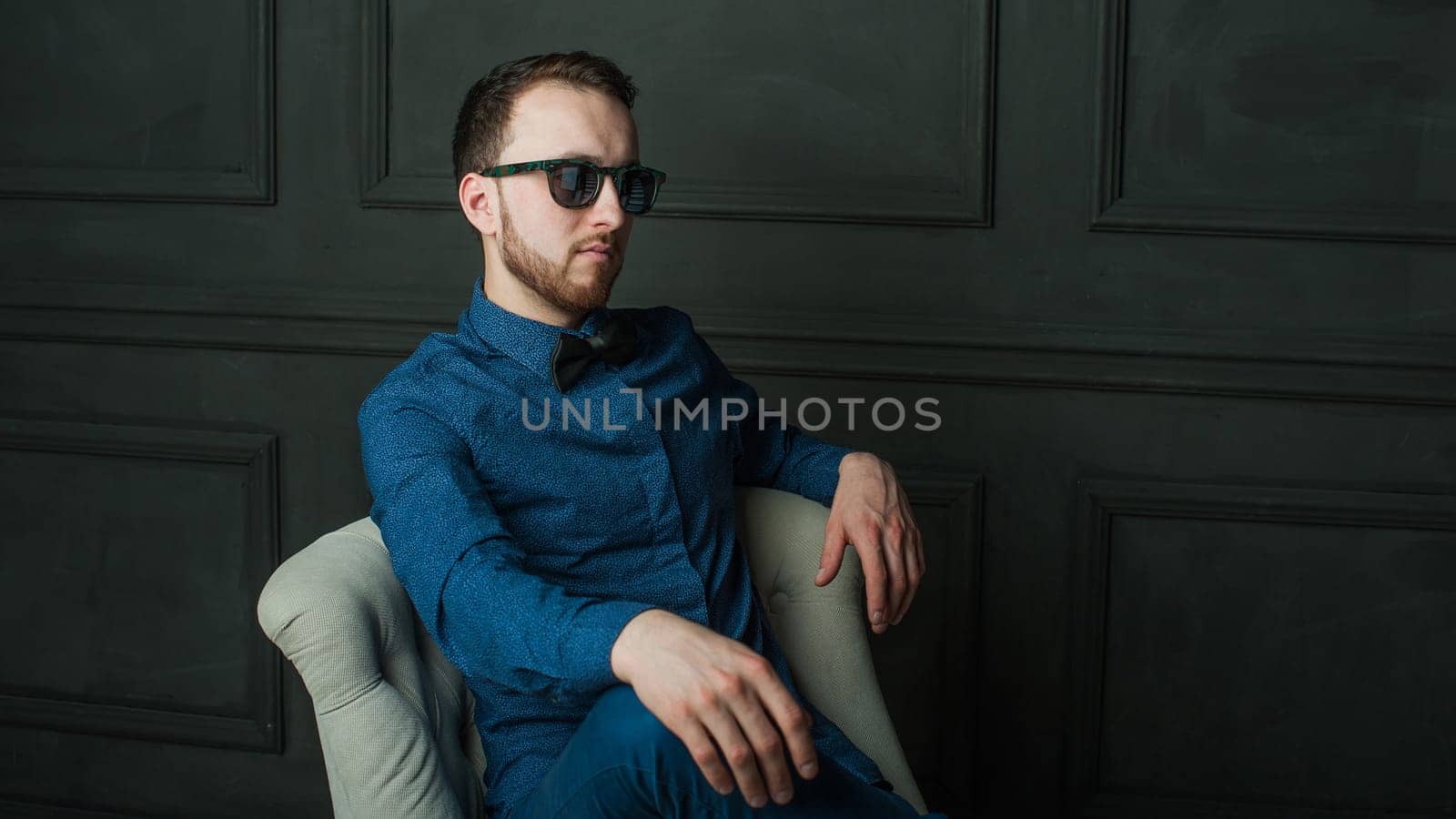 studio portrait of a young man on a dark wall background in a blue shirt and jeans. hipster man, dressed stylishly, with a beard and sunglasses, sitting in a light easy chair as a boss