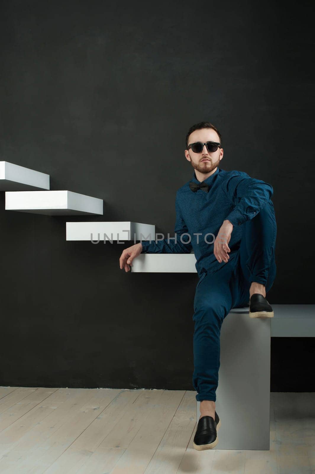 studio portrait of a young man on a dark wall background in a blue shirt and jeans. hipster man, dressed stylishly, with a beard and glasses, sitting on white stairs