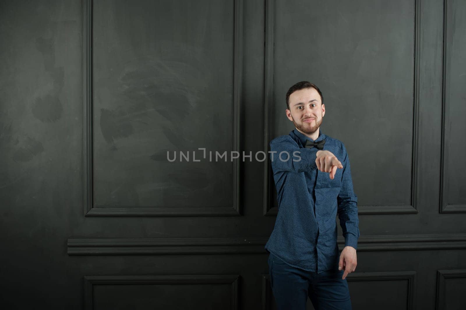 studio portrait of a young man on a dark wall background in a blue shirt and jeans. hipster man, dressed stylishly, with a beard, smiling and pointing finger