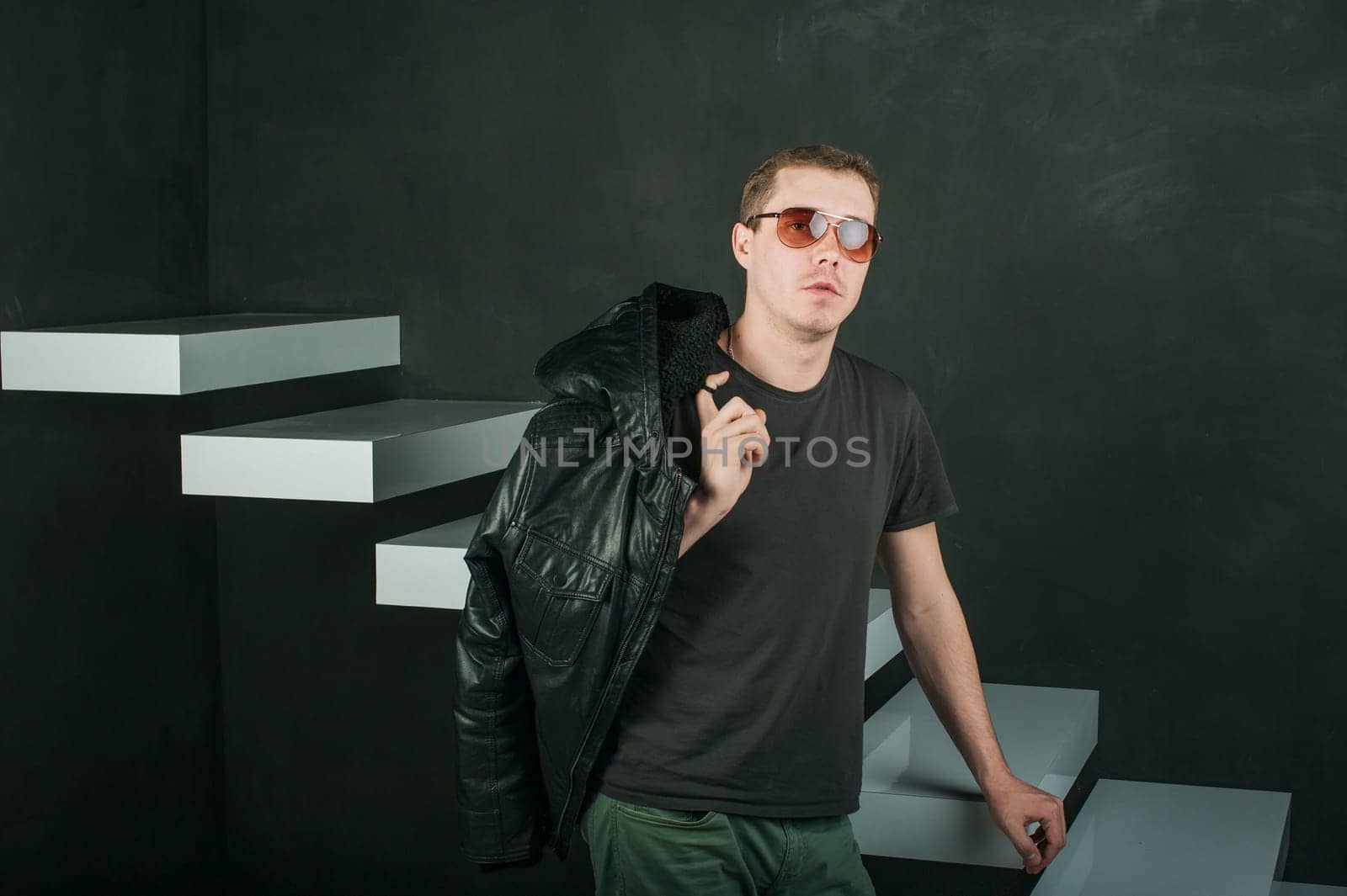 studio photography young brutal guy. man in sunglasses, T-shirt, jeans keeps black leather jacket, slung over his shoulder. black background painted walls and white stairs
