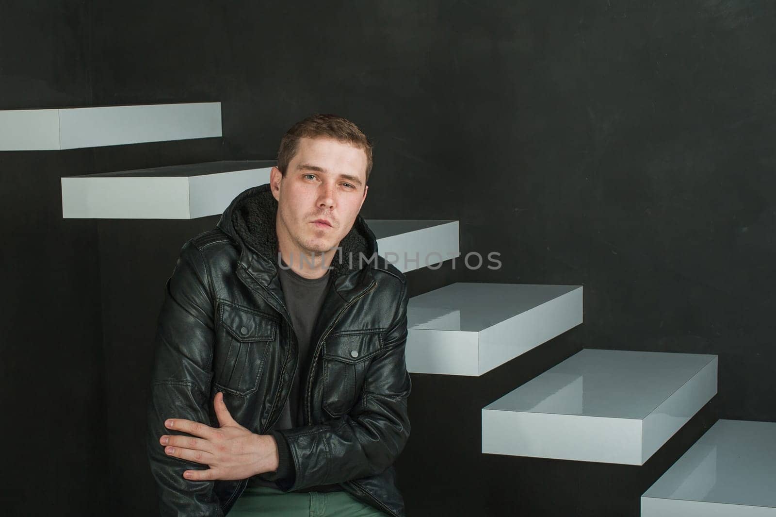studio photography of a young man in a dark T-shirt, black leather jacket. man sitting looking at the camera with arms crossed against a background of dark walls and white stairs