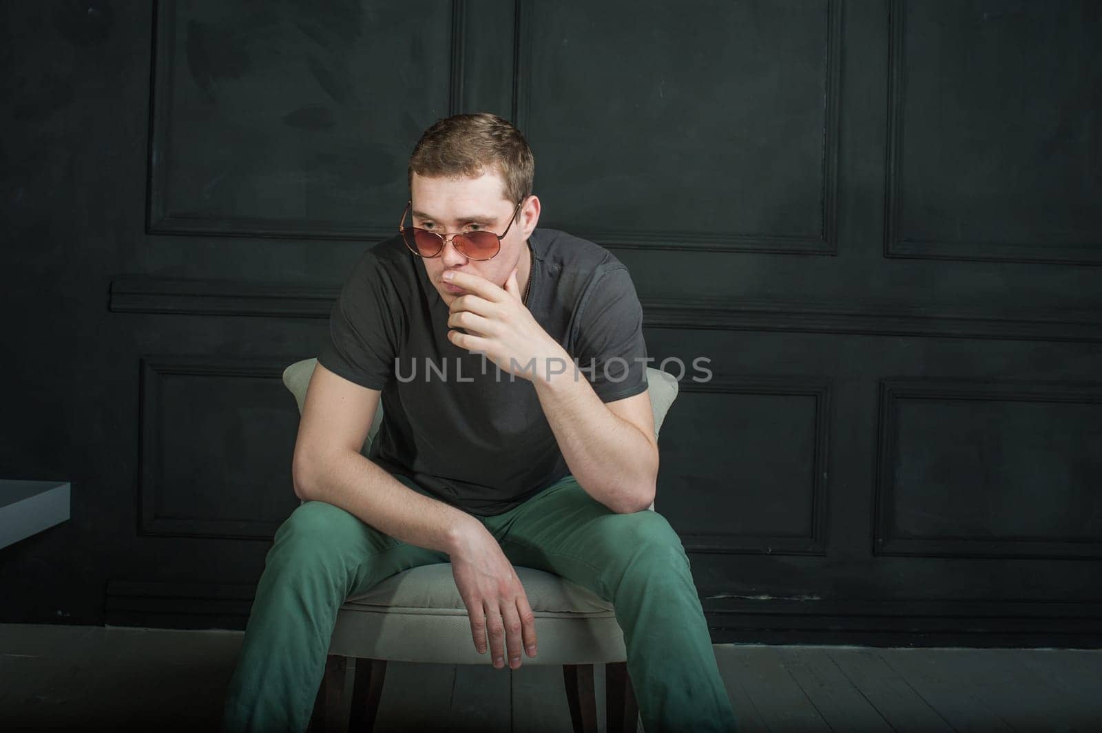 studio photography young brutal guy. man in sunglasses, T-shirt, jeans and high boots on a background of black wall, He is sitting in a white armchair throne.