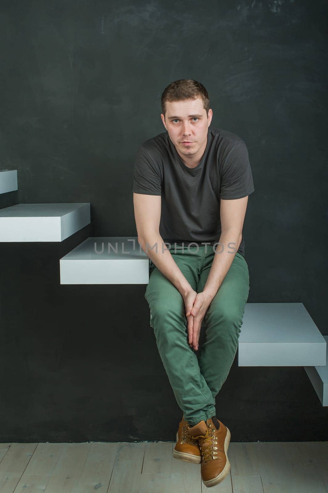 studio photography young brutal guy. man in T-shirt, jeans and high boots on a background of black wall on the white steps. sitting on the steps, his hands clasped palm to palm, looking at the camera