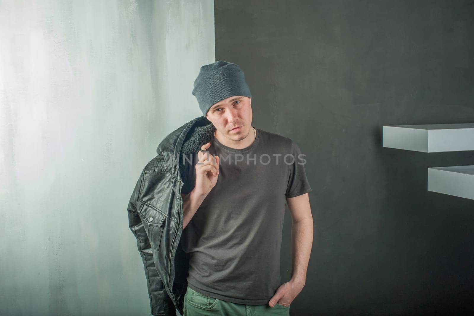 studio photography young brutal guy. man in T-shirt, jeans and a knitted cap, keeps black leather jacket, slung over his shoulder. black background painted walls and white stairs