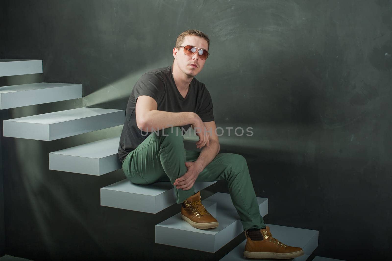 studio photography young brutal guy. man in sunglasses, T-shirt, jeans and high boots on a background of black wall on the white steps. He is looking at the camera while sitting on the stairs, hand on knee