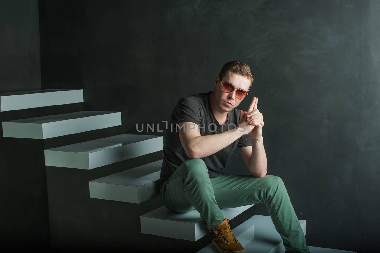 studio photography young brutal guy. man in sunglasses, T-shirt, jeans and high boots on a background of black wall on the white steps. sitting on the steps, holding hands in the shape of a gun for a shot