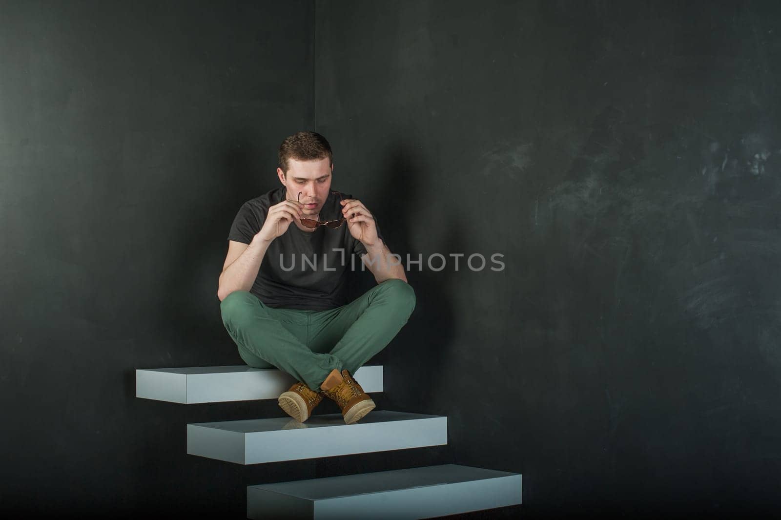 studio photography young brutal guy. man in sunglasses, T-shirt, jeans and high boots on a background of black wall on the white steps. sitting on the steps, took off his glasses and holds them in his hand
