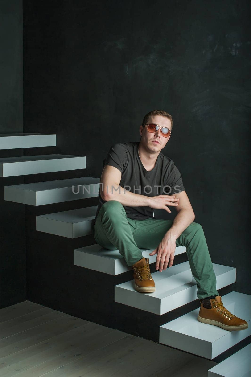 studio photography young brutal guy. man in sunglasses, T-shirt, jeans and high boots on a background of black wall on the white steps. He is looking at the camera while sitting on the stairs, hand on knee