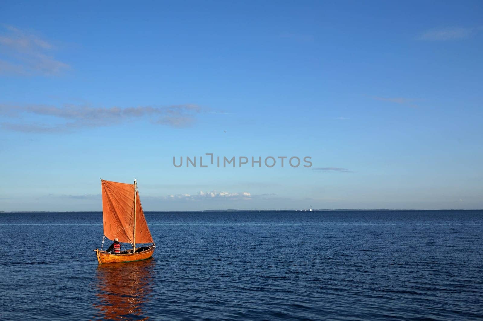 Small wooden boat with orange sail and anonymous person who controls the boat over the quiet ocean