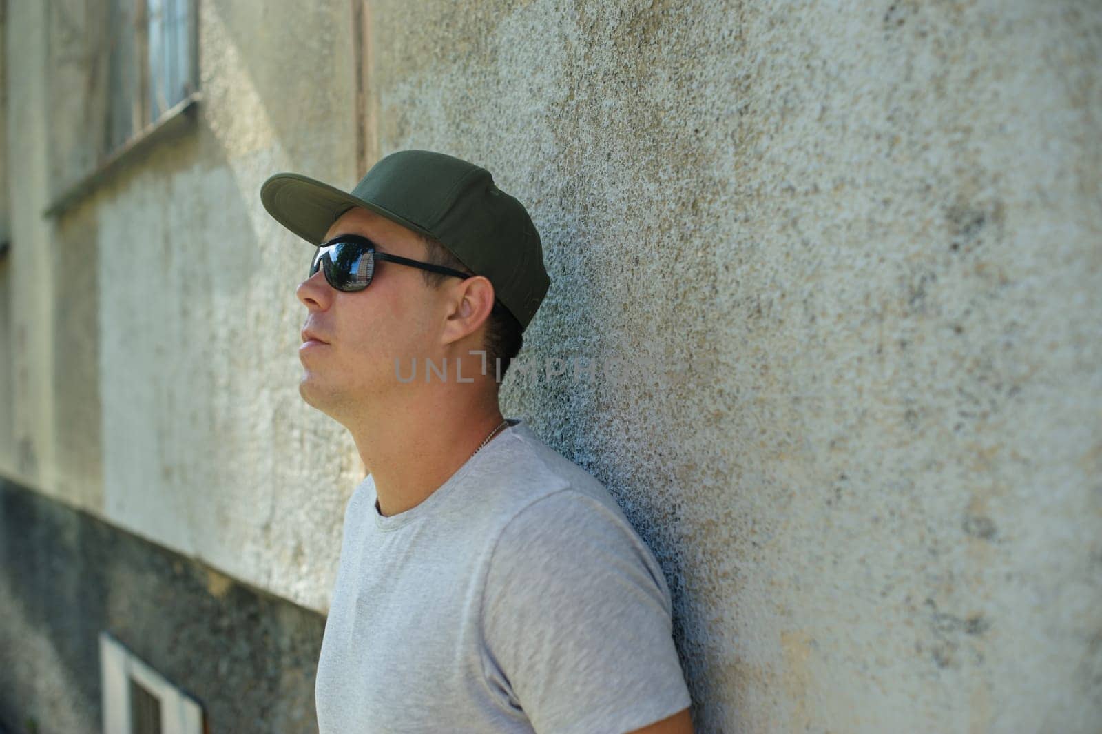 young guy in sunglasses standard physique posing on the street on a background of green bushes. He is wearing a light shirt and dark green cap. portrait of a man summer day