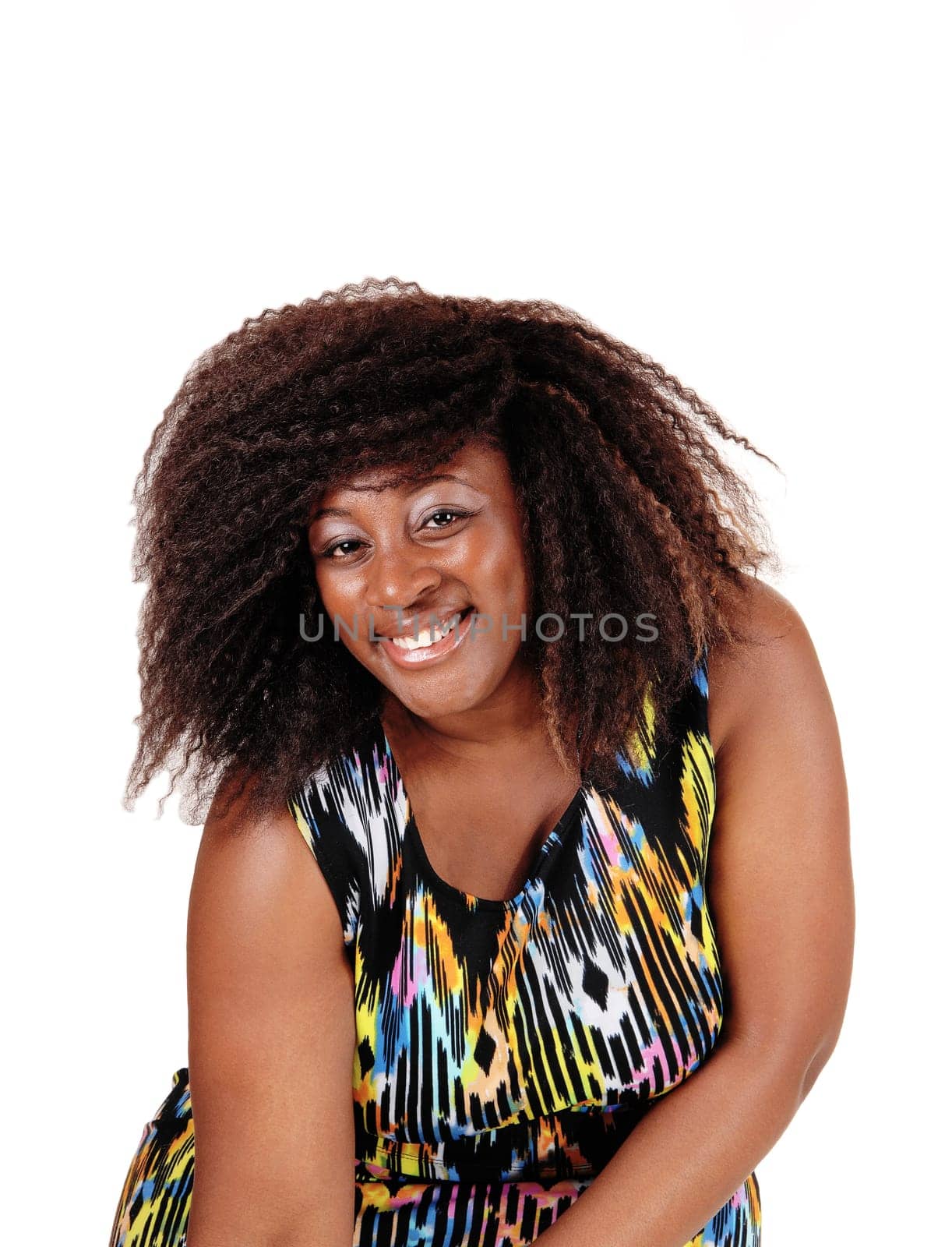A lovely smiling African American woman sitting in a colourful dress on a .chair, smiling into the camera, isolated for white background..