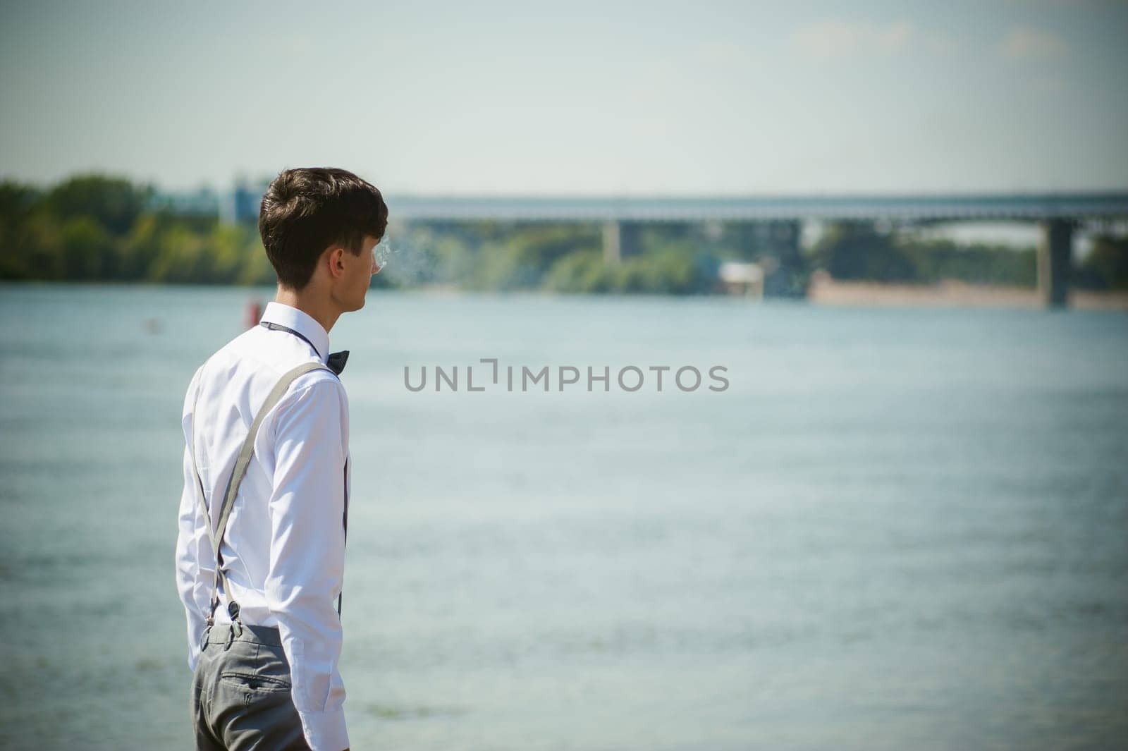 young skinny man, elegantly stylishly dressed in white shirt, trousers with suspenders and bow tie. vaper guy Smokes an e-cigarette, releas vapors of smoke, against background of river and bridge