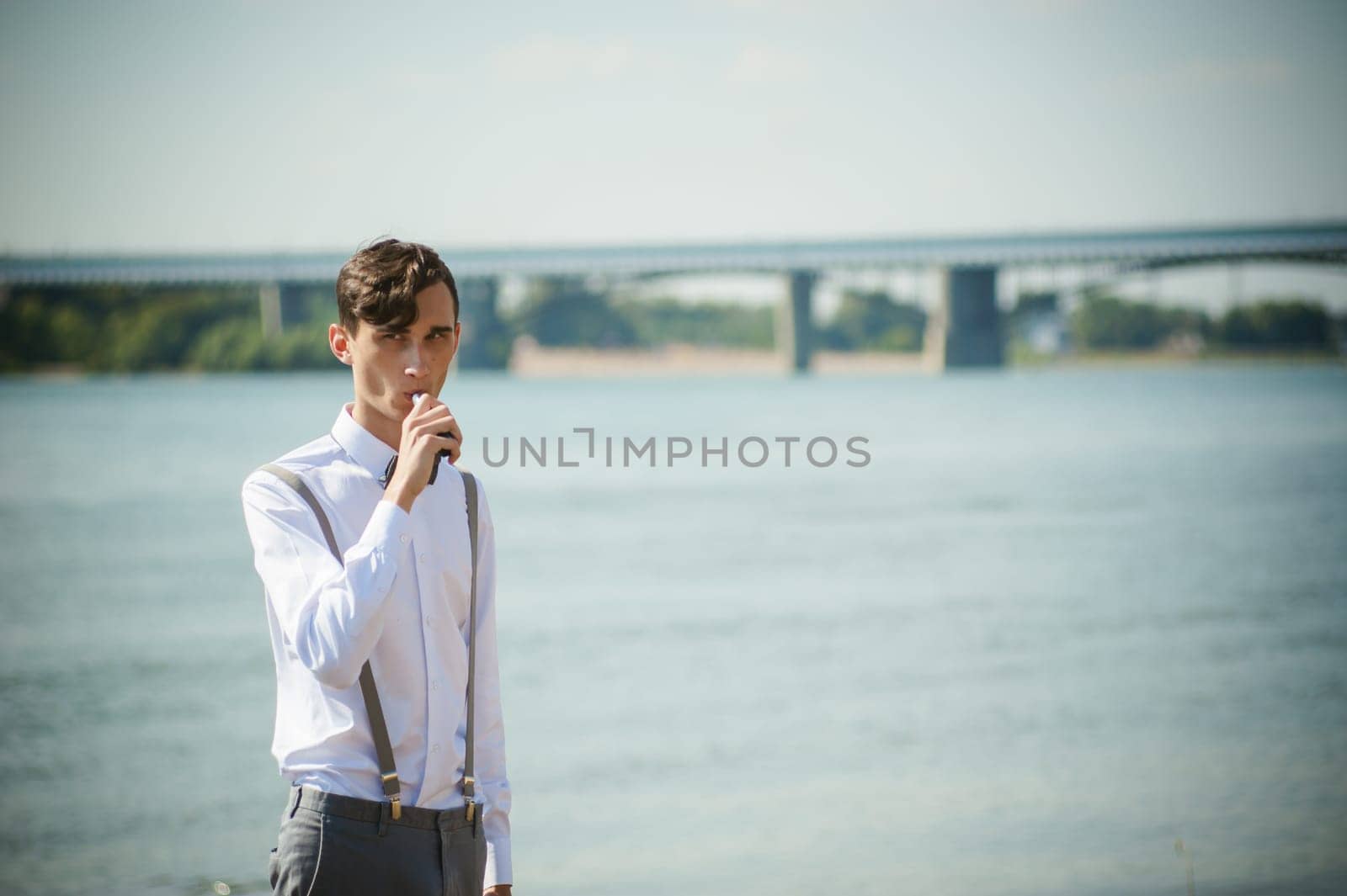young skinny man, elegantly stylishly dressed in white shirt, trousers with suspenders and bow tie. vaper guy Smokes an e-cigarette, releas vapors of smoke, against background of river and bridge
