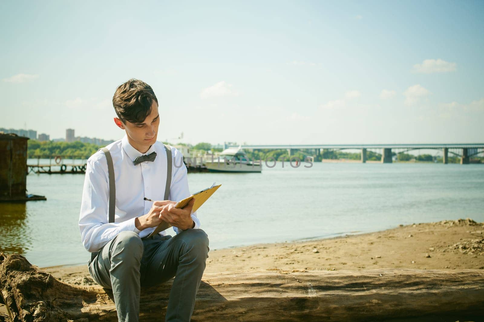 young skinny man, elegantly stylishly dressed in white shirt, gray trousers with suspenders and bow tie. young guy on pier on background of river and boats, takes notes of pen on sheet in notebook