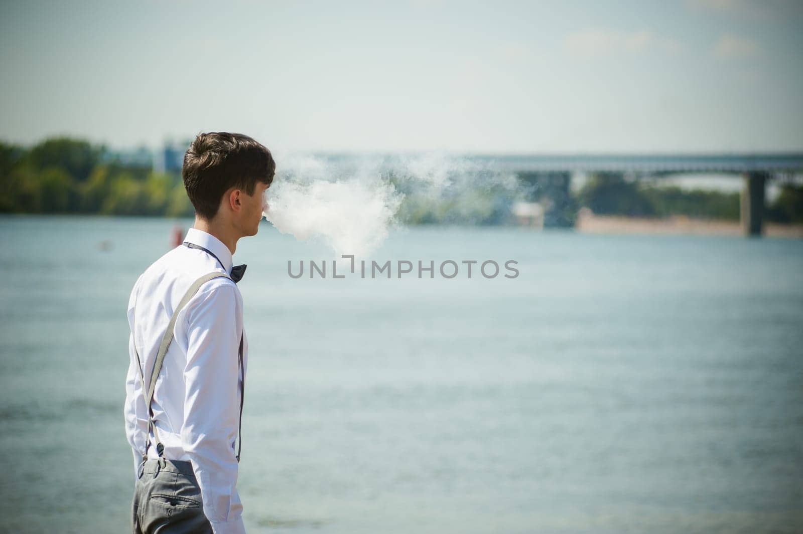 young skinny man, elegantly stylishly dressed in white shirt, trousers with suspenders and bow tie. vaper guy Smokes an e-cigarette, releas vapors of smoke, against background of river and bridge