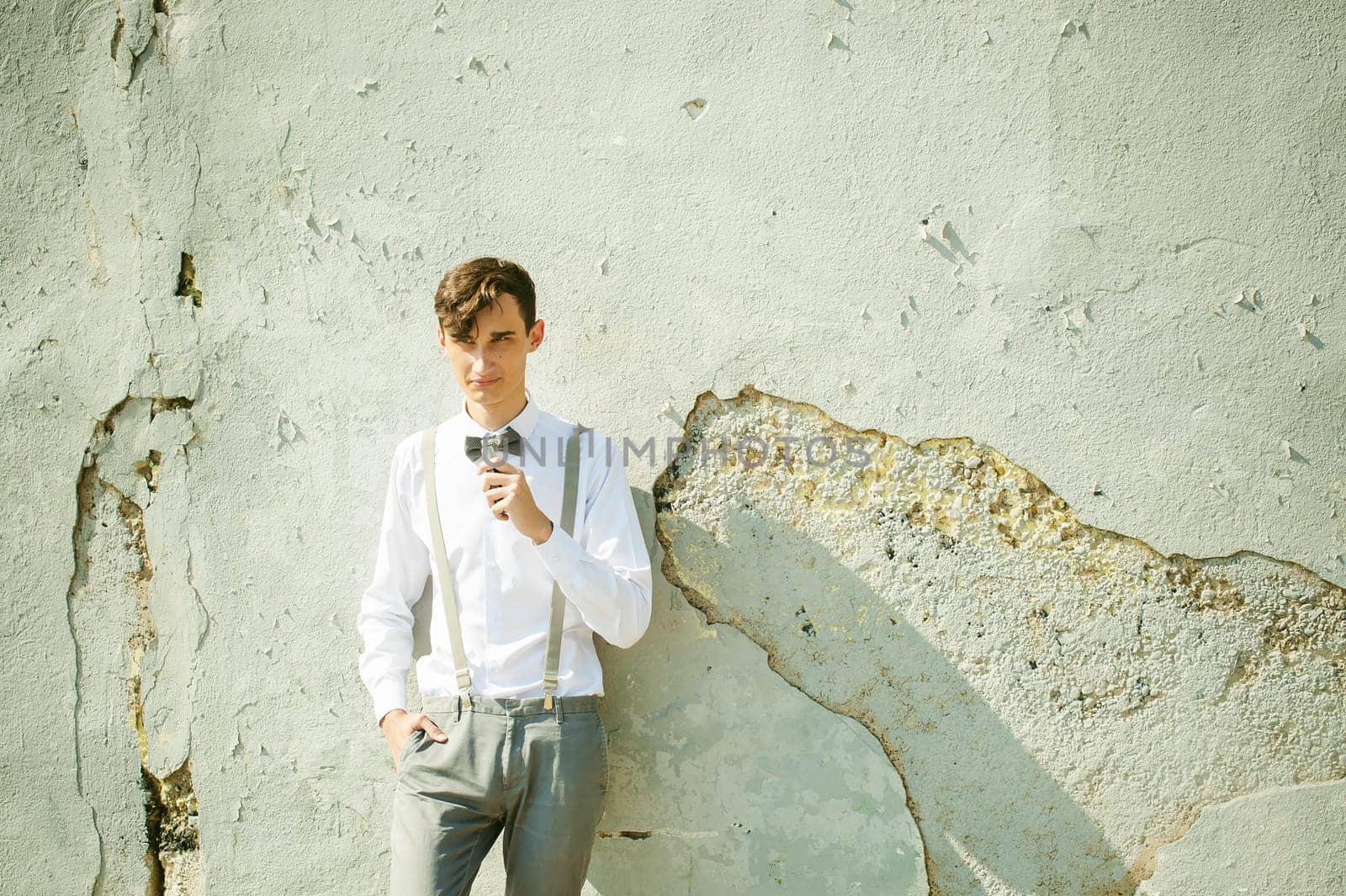 young skinny man, elegantly stylishly dressed in white shirt, gray trousers with suspenders and bow tie. Smokes an electronic cigarette on background of shabby wall, warm, sunny summer day
