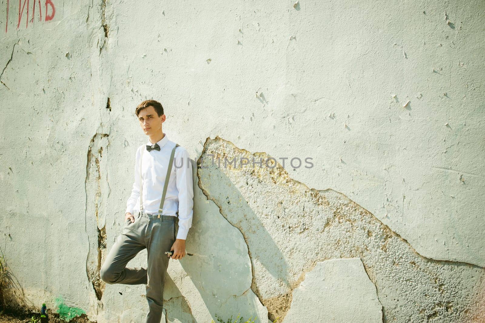 young skinny man, elegantly stylishly dressed in white shirt, gray trousers with suspenders and bow tie. Smokes an electronic cigarette on background of shabby wall, warm, sunny summer day
