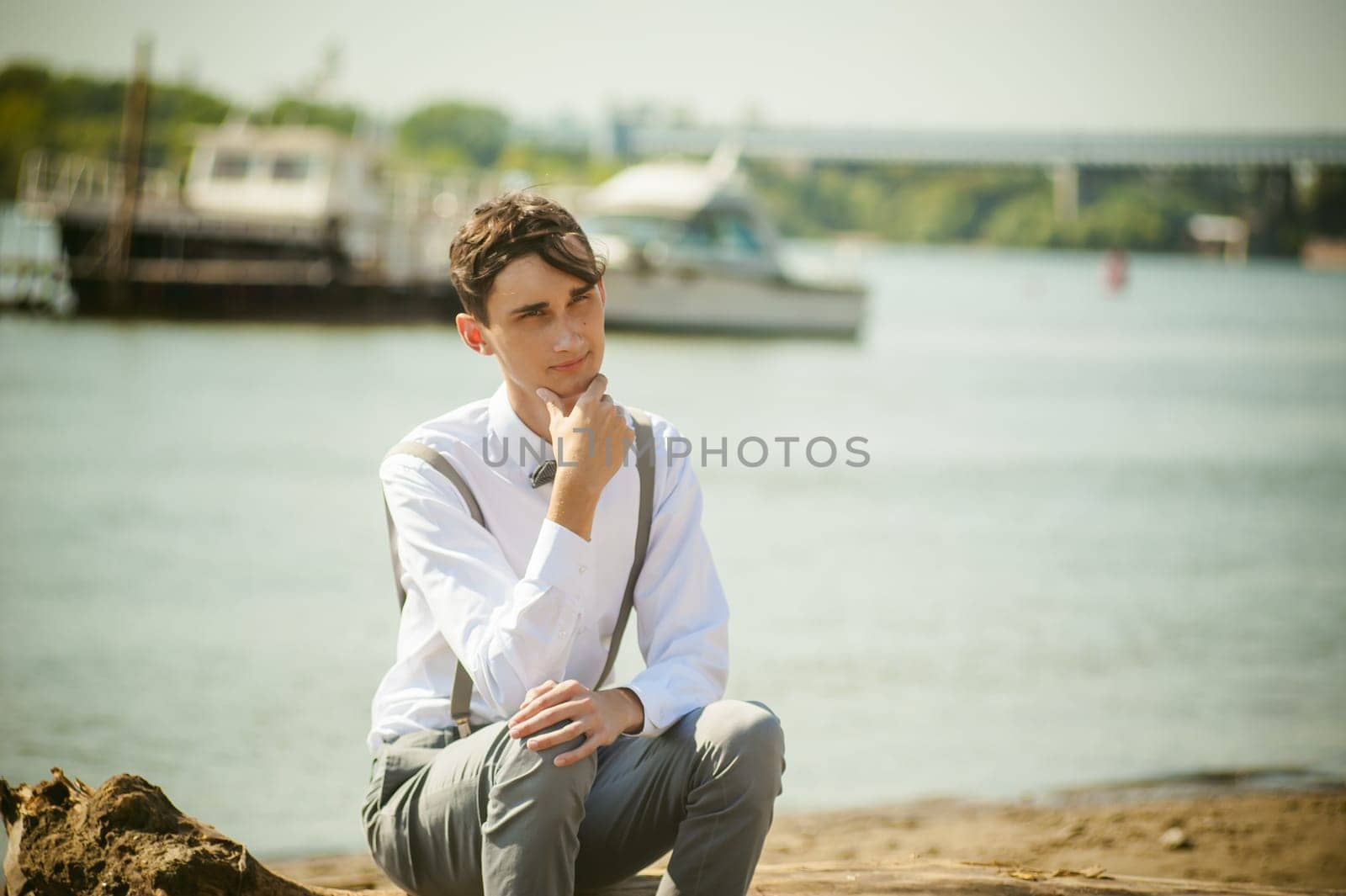 young skinny man, elegantly stylishly dressed in white shirt, gray trousers with suspenders and bow tie. Portrait young guy on pier on background of river and boats