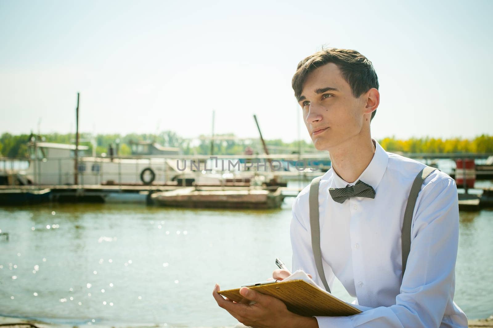 young skinny man, elegantly stylishly dressed in white shirt, gray trousers with suspenders and bow tie. young guy on pier on background of river and boats, takes notes of pen on sheet in notebook