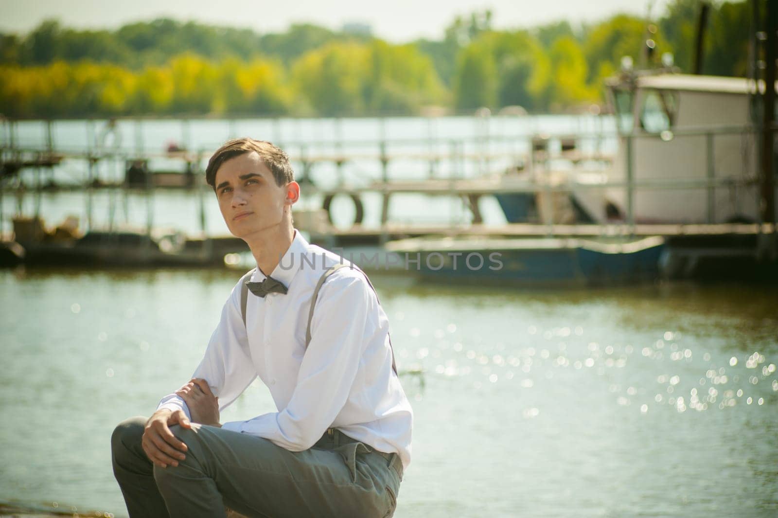 young skinny man, elegantly stylishly dressed in white shirt, gray trousers with suspenders and bow tie. Portrait young guy on pier on background of river and boats