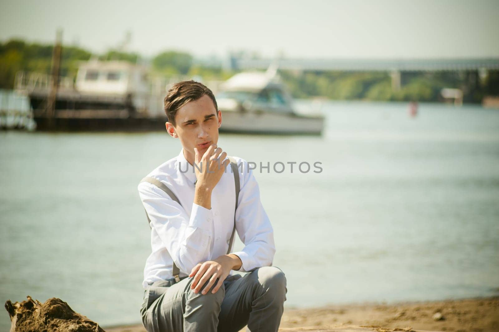 young skinny man, elegantly stylishly dressed in white shirt, gray trousers with suspenders and bow tie. Portrait young guy on pier on background of river and boats