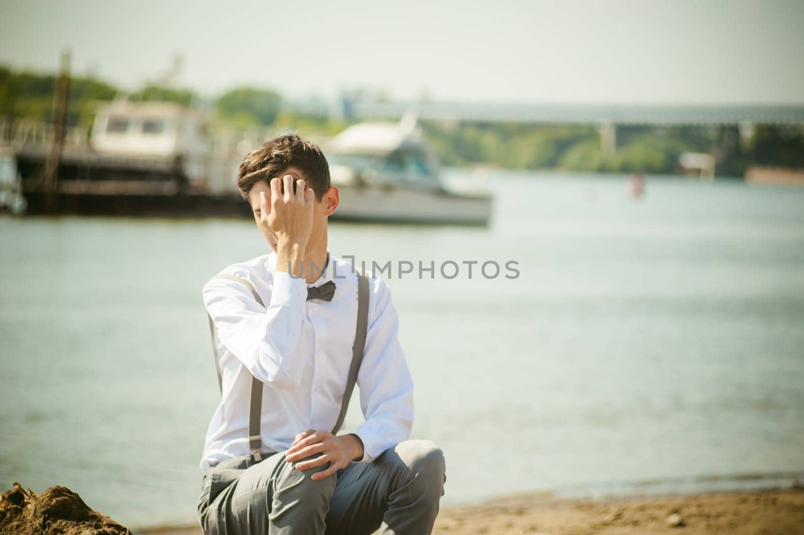young skinny man, elegantly stylishly dressed in white shirt, gray trousers with suspenders and bow tie. Portrait young guy on pier on background of river and boats