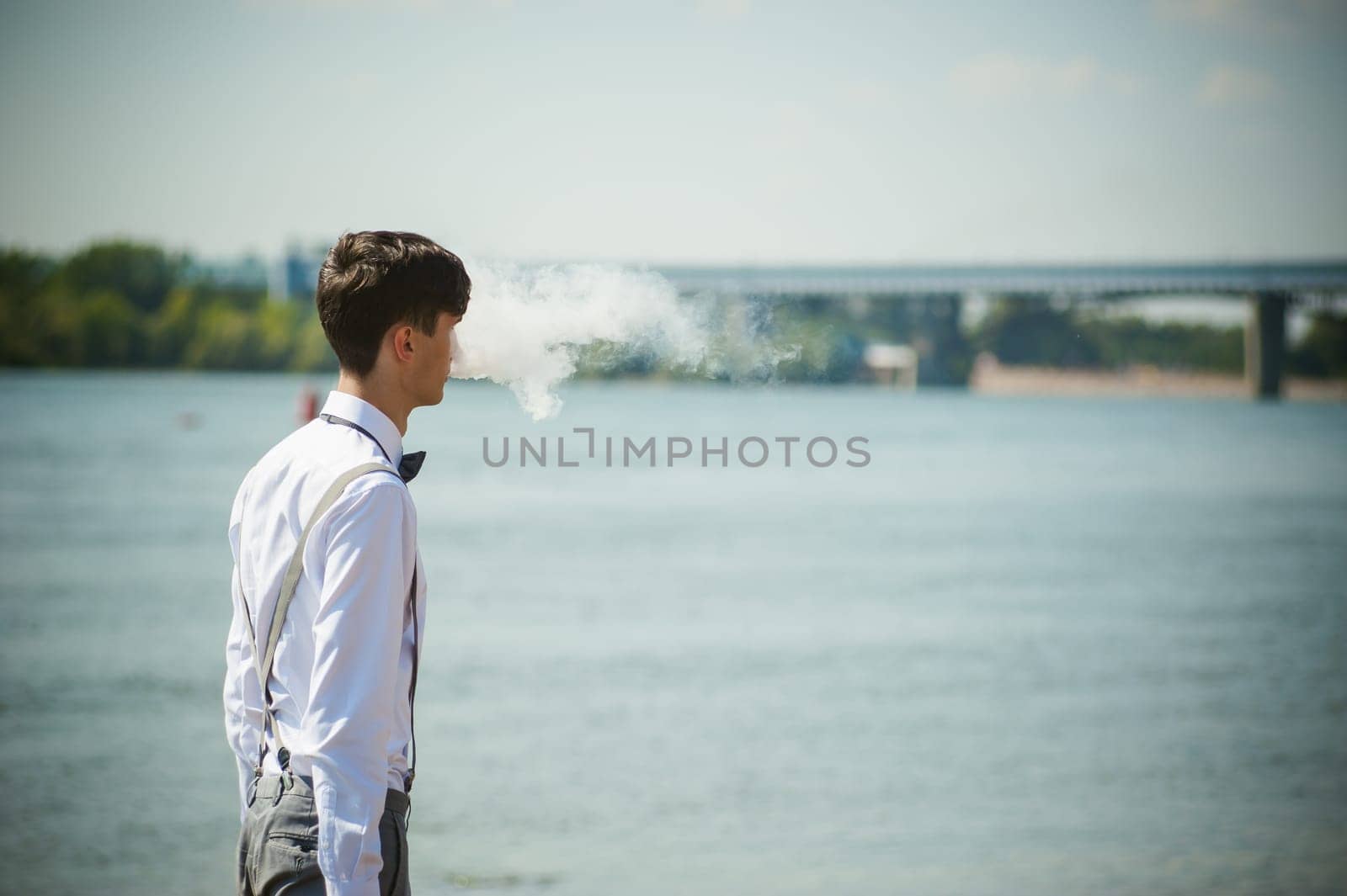 young skinny man, elegantly stylishly dressed in white shirt, trousers with suspenders and bow tie. vaper guy Smokes an e-cigarette, releas vapors of smoke, against background of river and bridge