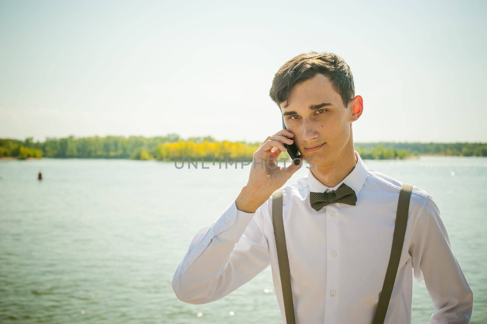 young skinny man, elegantly stylishly dressed in white shirt, gray trousers with suspenders and bow tie. Portrait of young guy talking on phone on background of river on warm sunny summer day