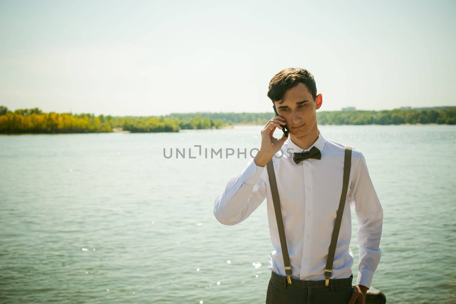 young skinny man, elegantly stylishly dressed in white shirt, gray trousers with suspenders and bow tie. Portrait of young guy talking on phone on background of river on warm sunny summer day