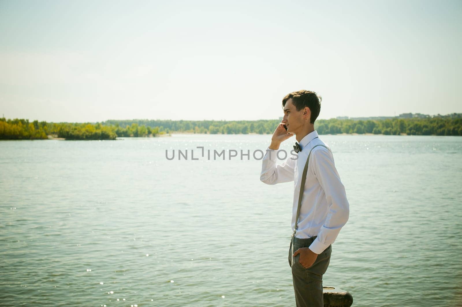 young skinny man, elegantly stylishly dressed in white shirt, gray trousers with suspenders and bow tie. Portrait of young guy talking on phone on background of river on warm sunny summer day