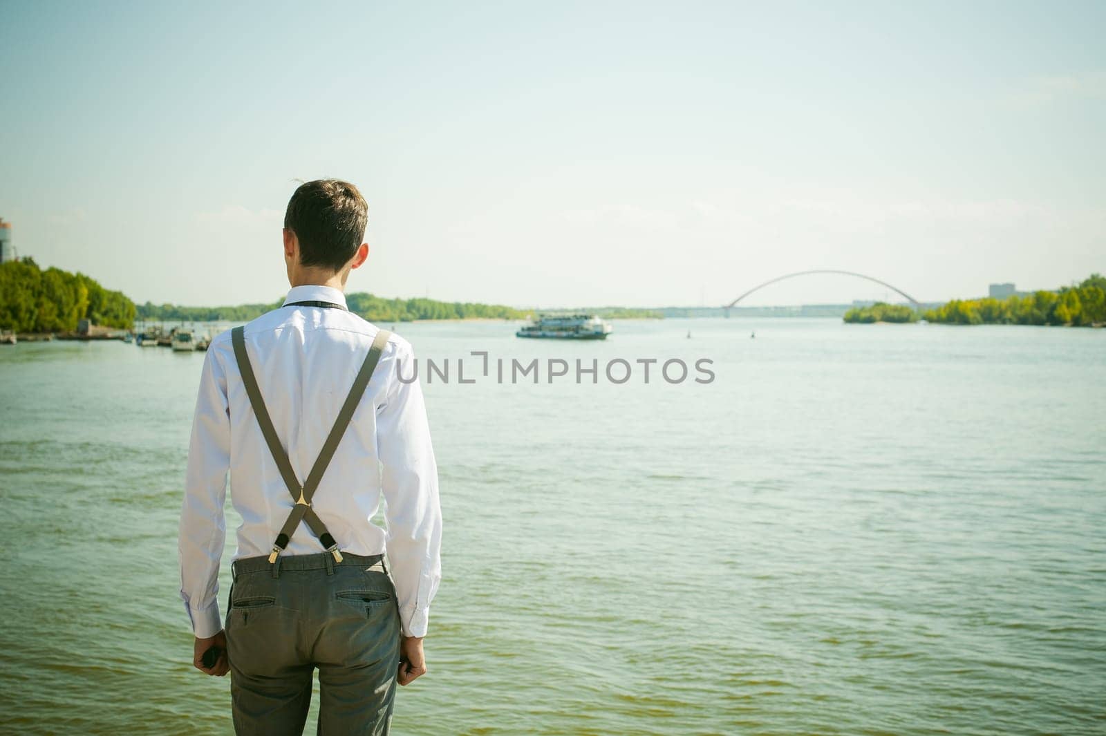 young skinny man, elegantly stylishly dressed in white shirt, gray trousers with suspenders and bow tie. Looking away, at the river water bridge and ship