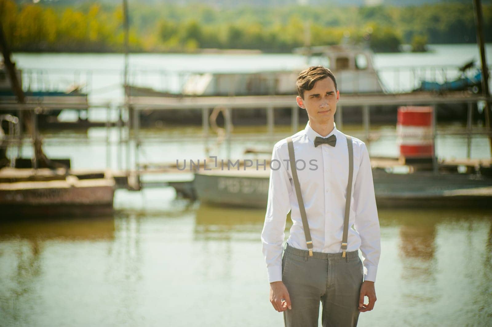 young skinny man, elegantly stylishly dressed in white shirt, gray trousers with suspenders and bow tie. Portrait young guy on pier on background of river and boats