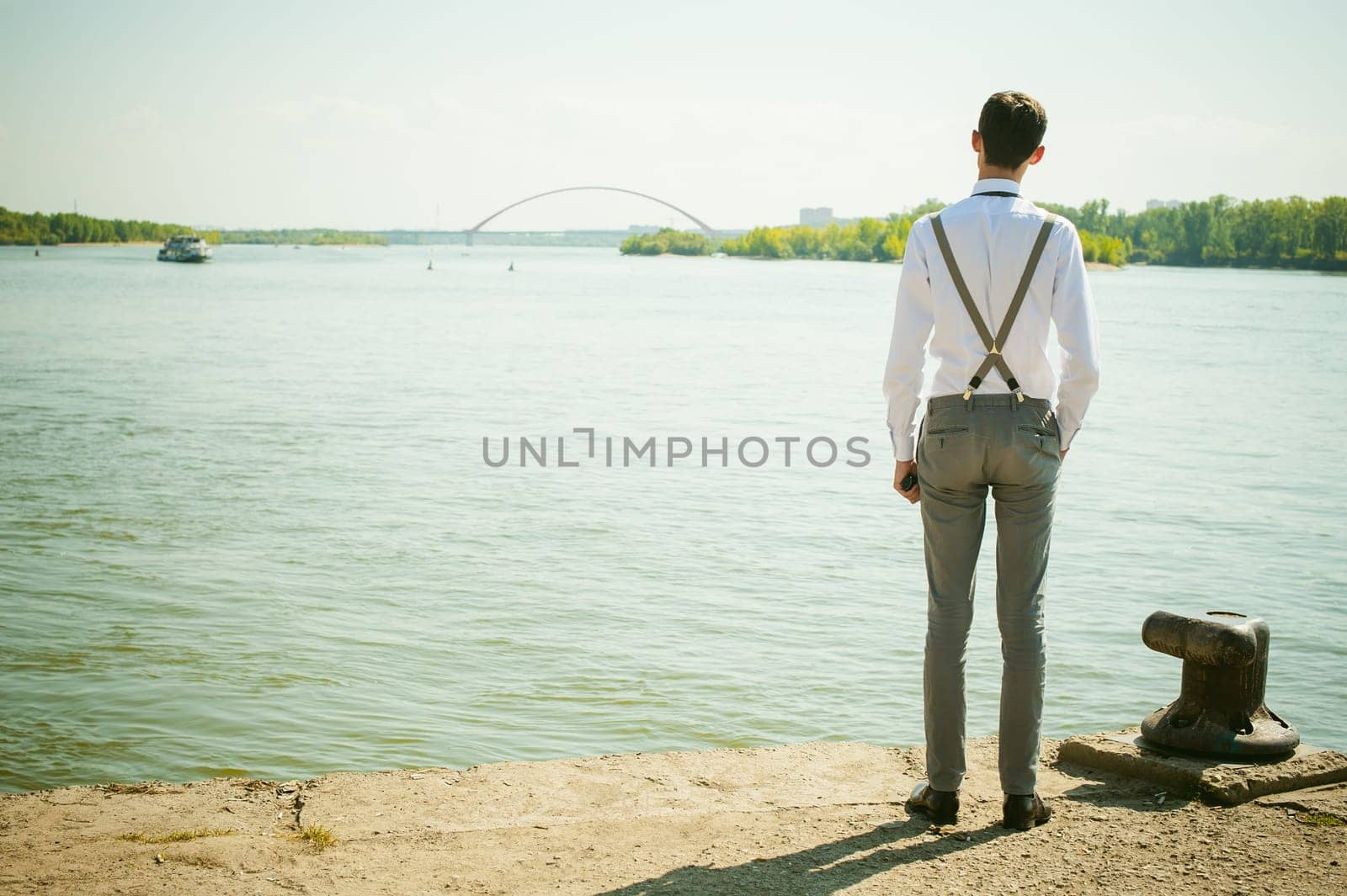 young skinny man, elegantly stylishly dressed in white shirt, gray trousers with suspenders and bow tie. Looking away, at the river water bridge and ship