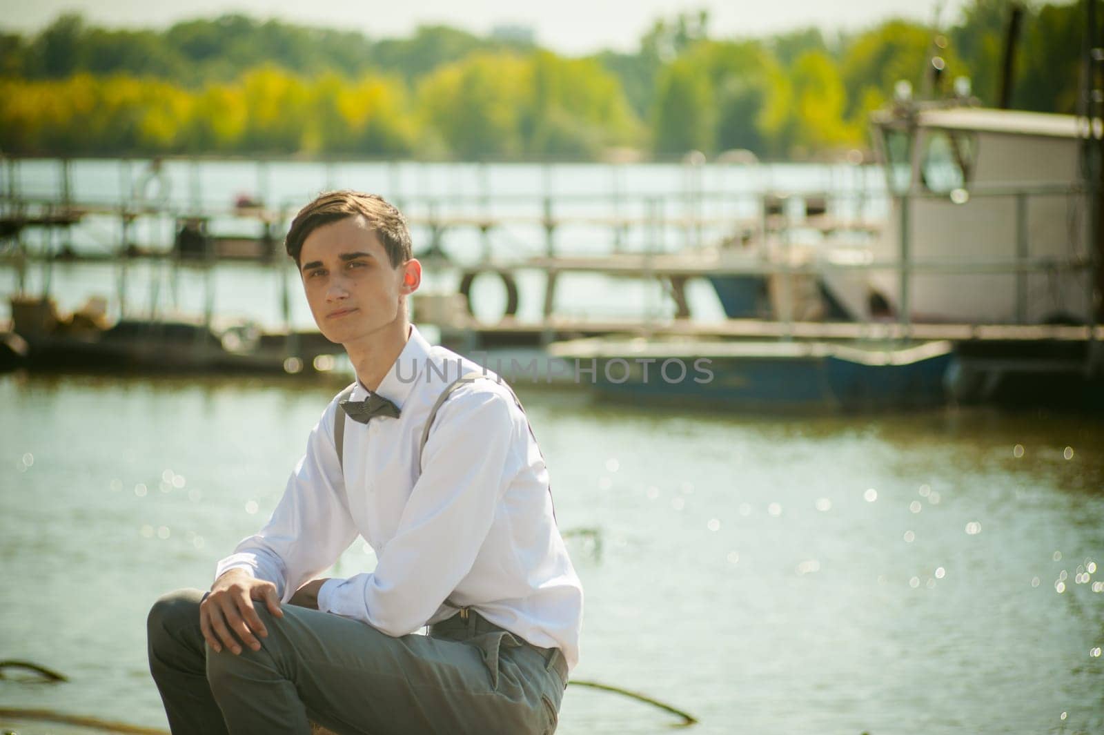 young skinny man, elegantly stylishly dressed in white shirt, gray trousers with suspenders and bow tie. Portrait young guy on pier on background of river and boats