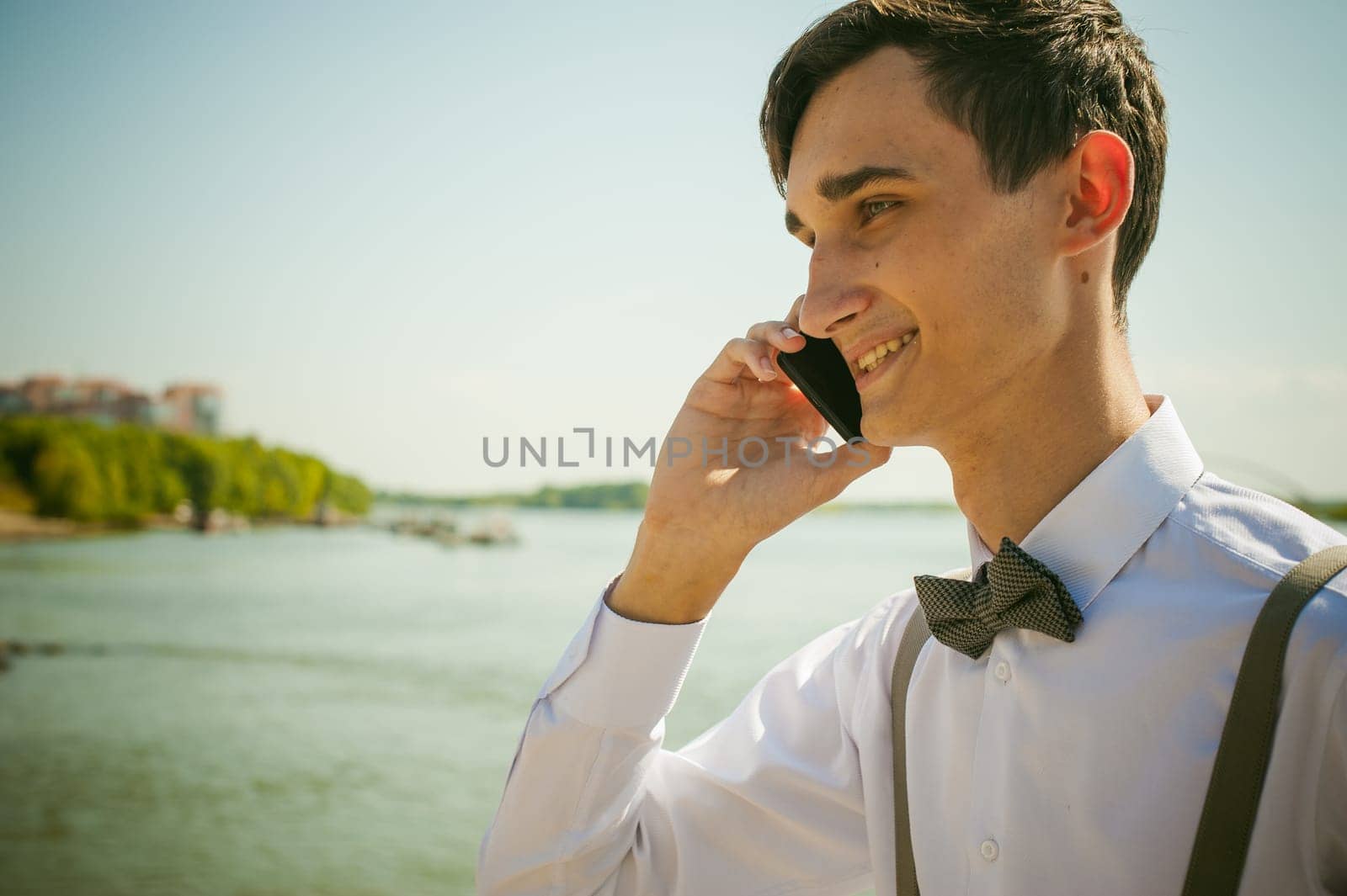 young skinny man, elegantly stylishly dressed in white shirt, gray trousers with suspenders and bow tie. Portrait of young guy talking on phone on background of river on warm sunny summer day
