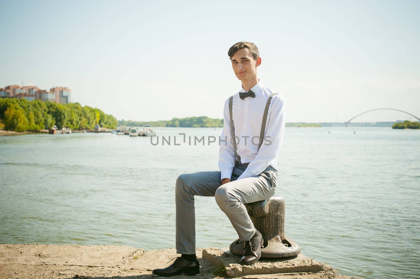 young skinny man, elegantly stylishly dressed in white shirt, gray trousers with suspenders and bow tie. guy sitting on a pier in the background of the river