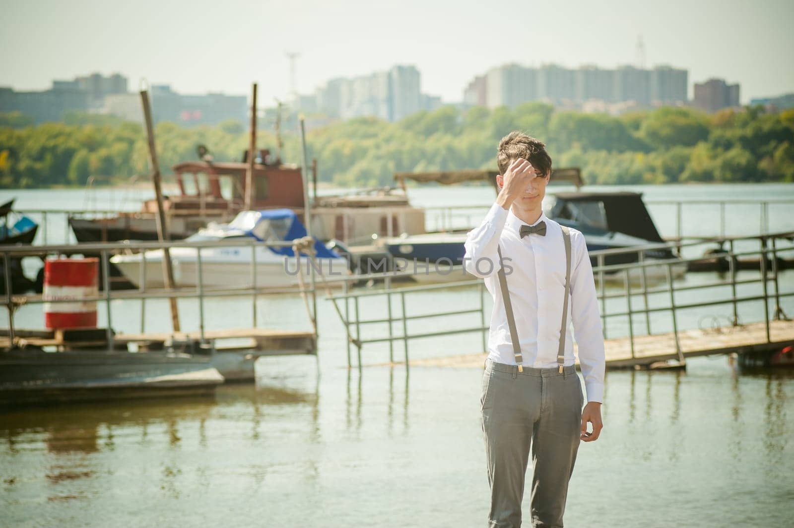 young skinny man, elegantly stylishly dressed in white shirt, gray trousers with suspenders and bow tie. Portrait young guy on pier on background of river and boats